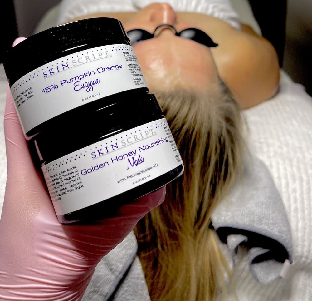 A gloved hand holding Skin Script Pumpkin-Orange Enzyme and Golden Honey Nourishing Mask jars over a client